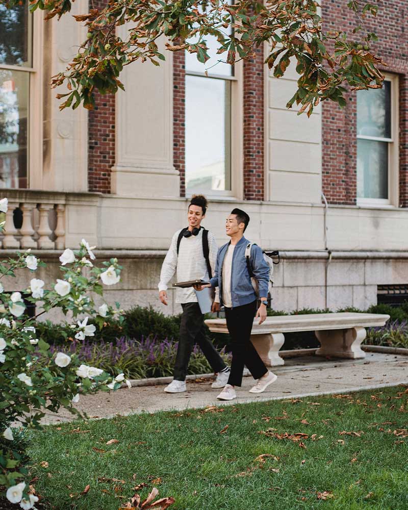 Two students walking along a path on a college campus with manicured lawn and plants